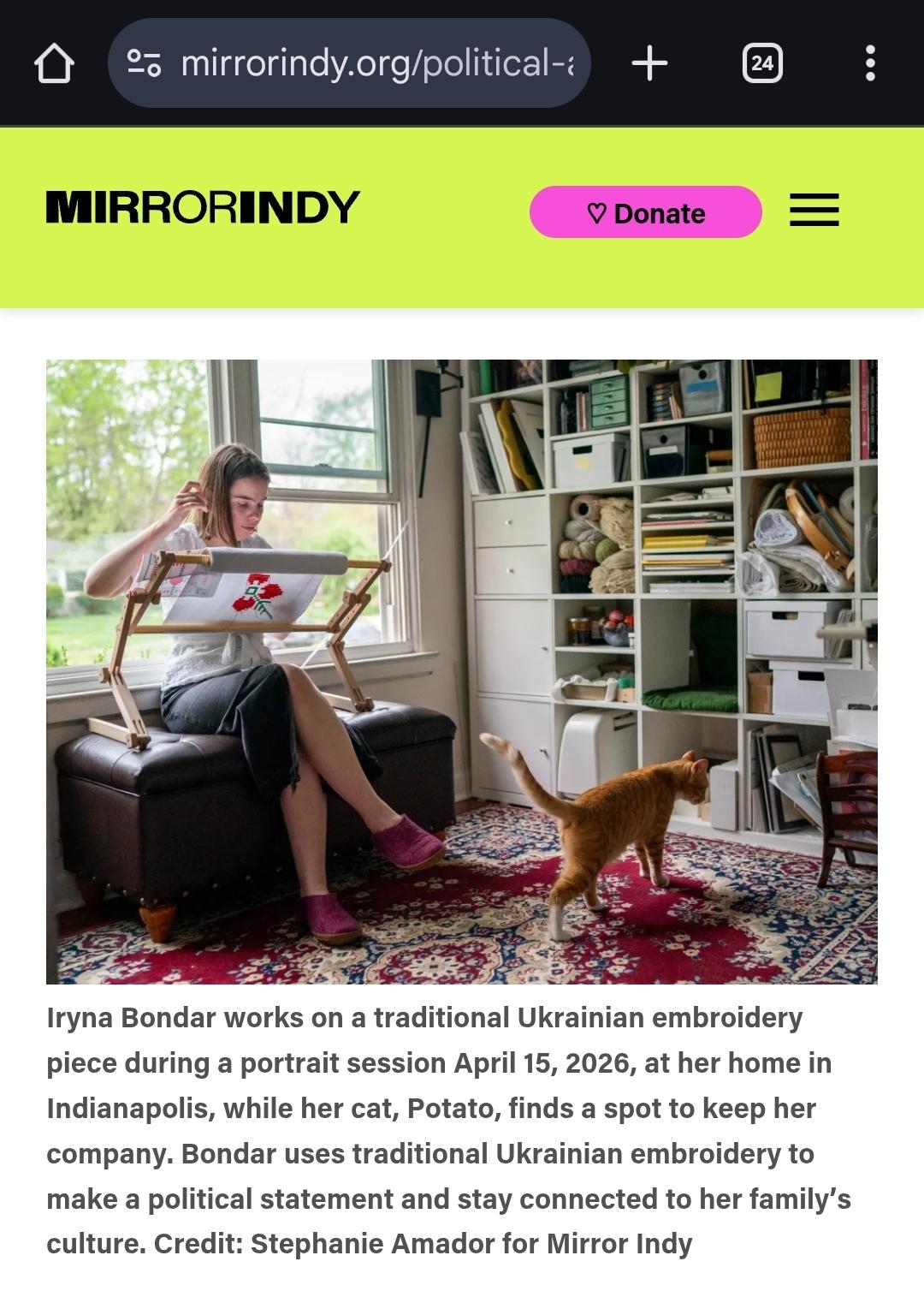Screenshot of Mirror Indy article: image with a woman sitting crosslegged, embroidering on fabric loaded in a frame, to her right is a big cube shelf of craft supplies, and on the floor an orange cat looking for a spot for itself to lay down.

The text below says:
Iryna Bondar works on a traditional Ukrainian embroidery piece during a portrait session April 15, 2026, at her home in Indianapolis, while her cat, Potato, finds a spot to keep her company. Bondar uses traditional Ukrainian embroidery to make a political statement and stay connected to her family’s culture. Credit: Stephanie Amador for Mirror Indy